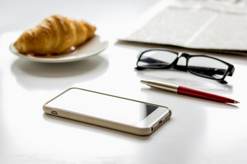 Office breakfast with croissant, phone and newspaper on white desk