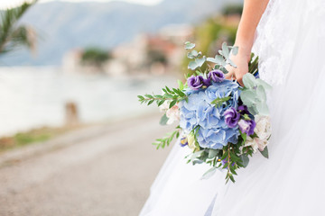 Bride holds wedding bouquet from blue flowers hydrangea and stands on light background of sea on...