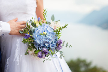 Bride holds wedding bouquet from blue flowers hydrangea and stands on light background of sea on...