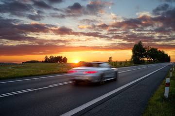 Speeding motion blur white car on the road in a rural landscape at sunset.