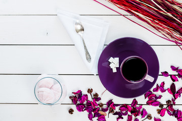 cup of tea or coffee on violet plate, silver spoon, marshmallow in glass vase, lump sugar, violet purple dry decor scattered on white colored wooden table,  top view