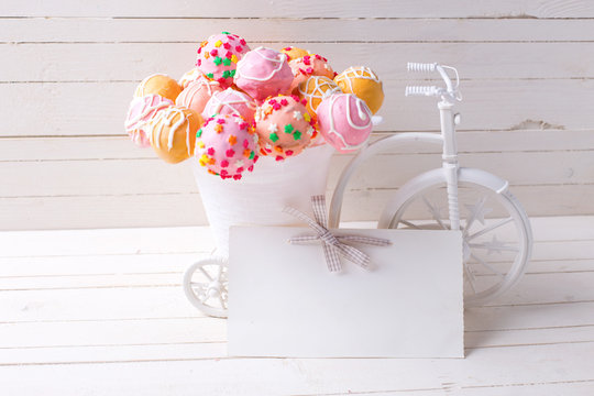Empty tag and bright cake pops  in decorative bicycle on white wooden background.