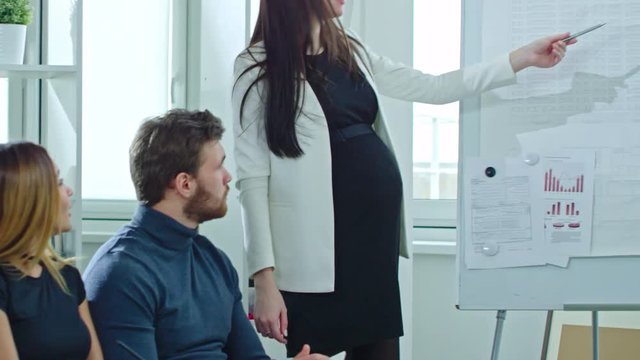  Panning shot of pregnant businesswoman and bearded businessman standing before whiteboard and explaining data on current company finances to coworkers 