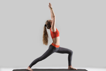 Young attractive yogi woman practicing yoga, standing in Warrior one exercise, Virabhadrasana I pose, working out, wearing sportswear, red sports bra, pants, indoor full length, isolated, grey studio