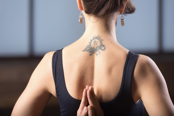 Fototapeta na wymiar Young attractive woman practicing yoga, making namaste behind the back, working out, wearing sportswear, black tank top, rear view, tattoo on her back, nice earrings, close up. Rear view