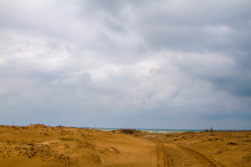 Photo of a sandy road to beach.  There is only sand until the sea. Tyre trails are also visible. Most of the photo is covered with clouds.