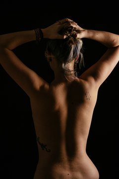 Chica mostrando  la espalda con tatuajes y con los brazos levantados. Foto de estudio con fondo negro.