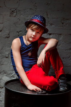 Portrait of cool young hip hop boy in blue hat and red pants sitting on black barrel in the loft