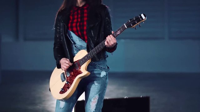 Cropped clip of young incognito woman in leather jacket and ripped jeans playing electric guitar and dancing in empty hall. Camera moving around