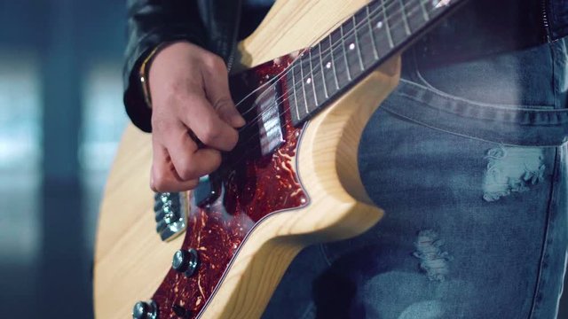 Cropped clip close shot of young incognito woman in leather jacket and ripped jeans playing electric guitar and dancing in empty hall. Camera moving around