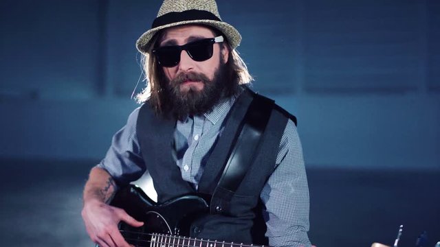 Close 4K shot of bearded man in sunglasses and hat playing the bass guitar sitting in smoke while video production process. Camera moving around