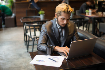 Young businessman holding cup of coffee while working on laptop computer in coffee shop.