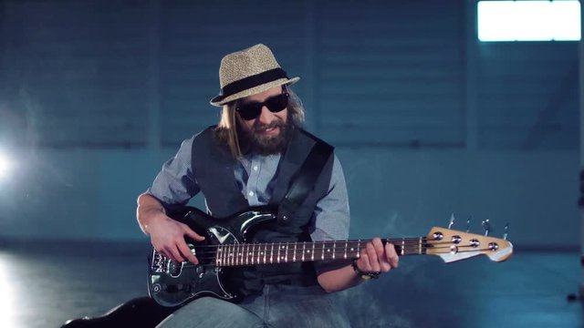 Bearded man in sunglasses and hat wearing vest and shirt sitting on chair, playing black electric guitar and smiling in empty hall with studio lighting. Camera moving around
