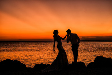 Silhouette of a loving, married couple where he is kissing her hand. In the background of the setting sun, rocks and sea.