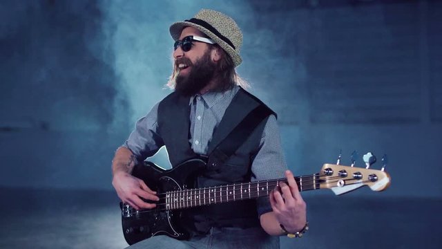 Bearded man in sunglasses and hat wearing vest and shirt sitting on chair, playing black electric guitar and smiling in empty hall with studio lighting. Camera moving around
