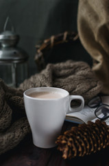 White cup of cocoa on an old wooden table with an entertaining book and reading glasses.