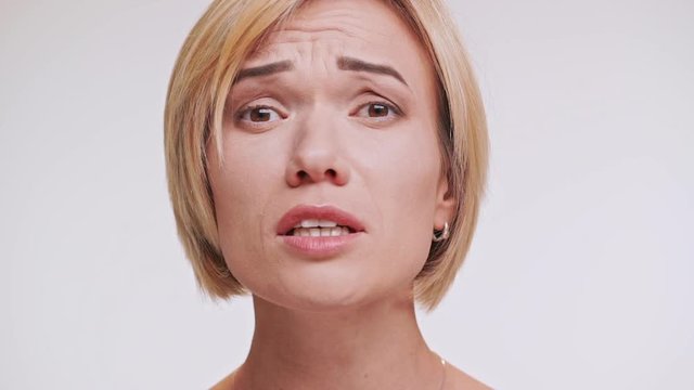 Confused middle-aged Caucasian woman with blonde hair looking at camera with upset. Close up footage on white background in slowmotion