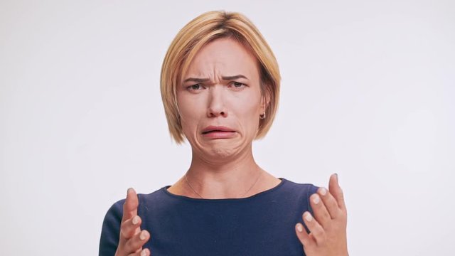 Middle-aged Caucasian female in blue sweatshirt and blonde hair looking at camera in disgust gesturing on white background in slowmotion