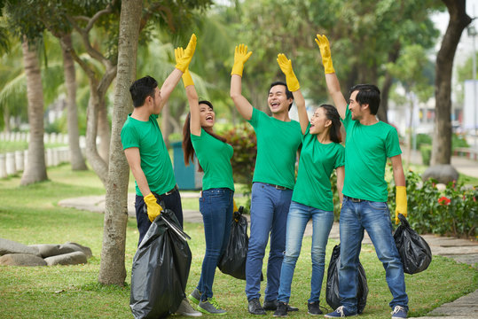 Group of enthusiastic young people holding full bin bags and raising hands as sign of successful completion of waste collection
