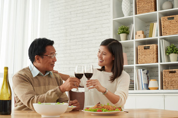 Senior Asian man and his young daughter celebrating Fathers Day together: they holding glasses of wine in hands and clinking them together
