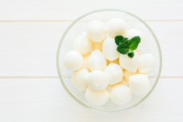 Small balls of mozzarella in glass bowl decorated fresh mint leaves on white wooden table. Selective focus