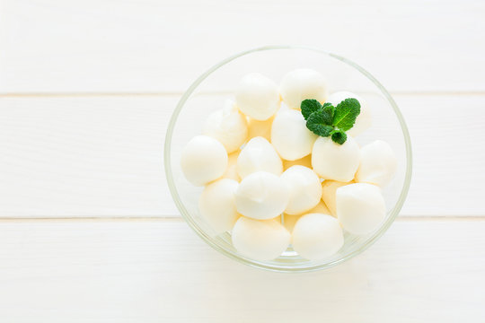 Small balls of mozzarella in glass bowl decorated fresh mint leaves on white wooden table. Selective focus