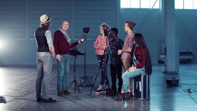 Group of young people filmmakers standing in empty hall with shooting gear and the director showing how to set lighting on actor, switching the studio light stand on