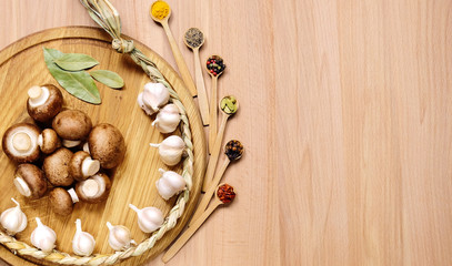 Braid of garlic, mushrooms and bay leaves on a round cutting board light wooden table. Nearby are several small wooden spoons with different spices. Top view.