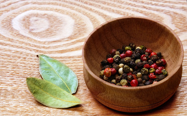 Fragrant colorful pepper in a wooden bowl and two dried bay leaves on a light wooden table.