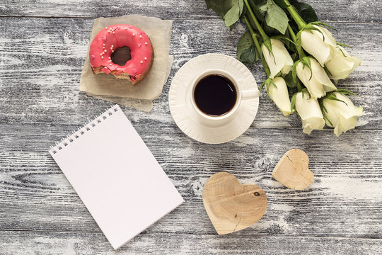 Blank notebook, cup of coffee, donut, hearts and white roses on a gray wooden table. Top view.