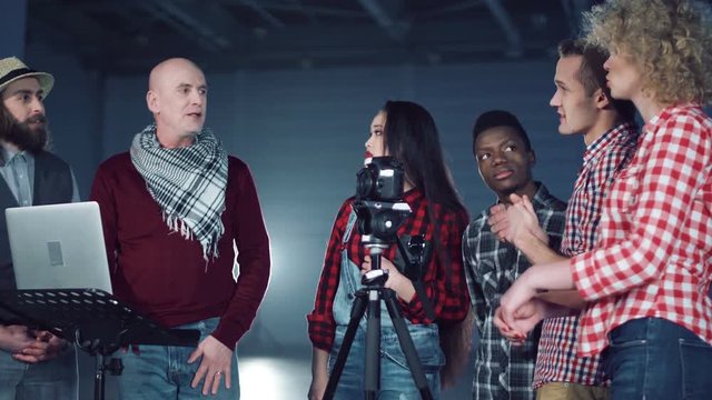 Bold man producer with laptop talking to crew of young mixed ethnic people filmmakers standing near DSLR camera on tripod in dark empty hall