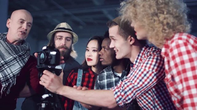 Bold man producer with laptop talking to crew of young mixed ethnic people filmmakers standing near DSLR camera on tripod in dark empty hall