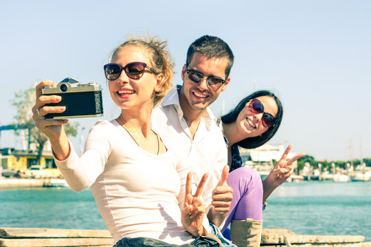 Happy best friends taking selfie with vintage camera at pier - Cheerful students on vacation having fun doing photo by the ocean - Concept of joyful moments with attractive playful teenagers outdoors