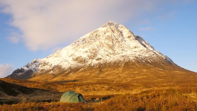 Green touristic tent on meadow at river below snowy cone of mountain Stob Dearg 1021 metres high. Higland in Scotland an marvelous sunny winter day. Dry grass and heather bushes. 