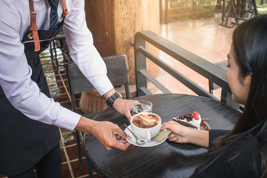Close up of man serving coffee while standing in coffee shop. Focus on male hands placing a cup of coffee on table. young waiter serving some coffee and a cupcake to a female customer in a cafe.