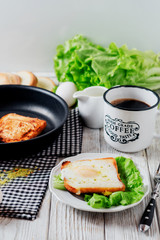 a delicious and hearty breakfast, fried eggs in a white toast with spices, peppers, lettuce, a cup of black coffee and milk on a wooden background 