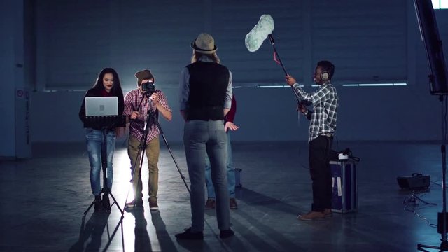 Group of young people with camera on tripod, laptop and boom mic setting gear for shooting scene with man in hat standing and playing role or giving interview
