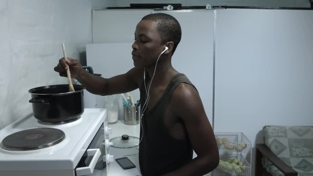 Young man listening and dancing to music on his mobile, while cooking at the stove.