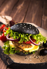 Delicious homemade vegan black burger with two chickpea cutlets, tomatoes, cheese, onion and salad on wooden table, dark background.