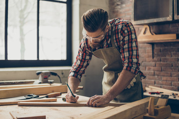 Bearded handsome cabinetmaker at the tabletop with pencil drawing sign on plank.  Stylish craftsman...