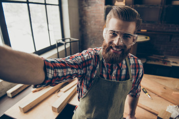 Happy successful handsome cabinetmaker make selfie photo. Stylish young entrepreneur with brutal hairstyle and glasses smiling and photographing at his workstation. Love job