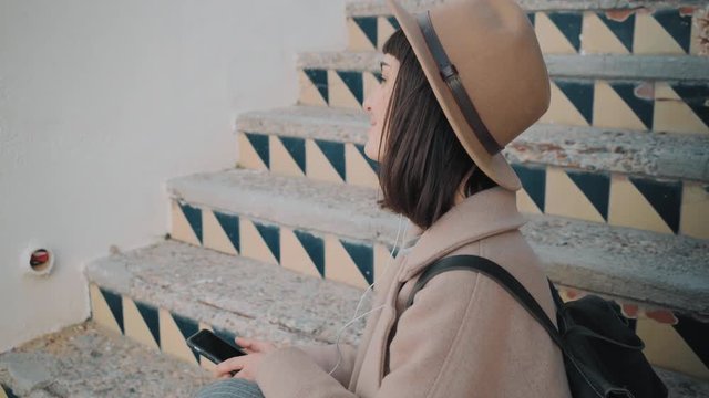 Camera moves around elegant girl sitting on old rustic stairs , listening her favorite music on smarphone, smiling and slightly moves on music tempo