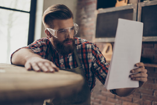 serious handsome  craftsman chek wood and document. Stylish young cabinet-maker with brutal hairstyle and saved glasses work at his workplace. He love his job