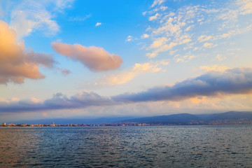 Beautiful morning seascape. Bright blue sky with cumulus clouds and a calm sea. The mountains and the city on the horizon. Dawn above the sea.