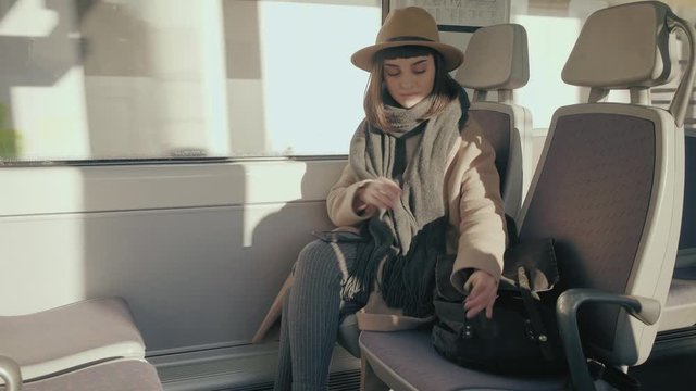Young lady in train looking for earbuds in her black rag backpack and connects headphones to smartphone, plugs buds in her ears and chooses music in playlist