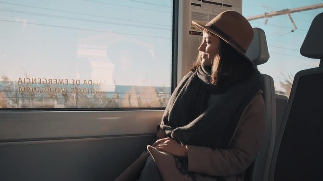 Attractive smiling fashionable girl travels in train at autumn or spring and takes photos or videos on her smartphone through window Sunny day