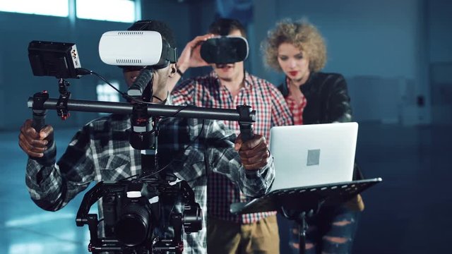 Young people testing camera gyro stabilizer gimbal with virtual reality headset on, looking around. Men and woman with laptop