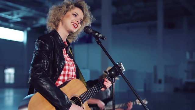 Young woman with curly hair wearing leather jacket and red checked shirt singing and playing guitar standing at microphone stand. Man band member with guitar sitting blurred in background