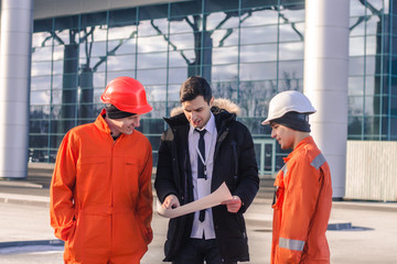 boss or Chief  instructs young team of  young engineers with a construction project. They wear overalls and safety helmets. Business modern background