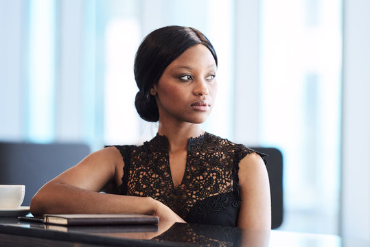 Beautiful Young Black Woman Sitting At A Modern Counter With One Elbow Resting On The Table And The Other Hanging Down, While Looking Off Into The Distance.